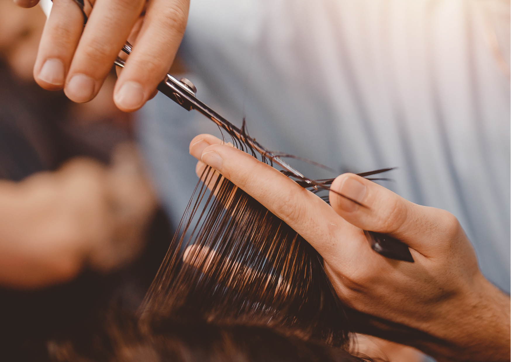 Em close, a fotografia mostra duas mãos de pessoa branca segurando mechas de um cabelo e usando pente e escova para realizar um corte nas pontas.