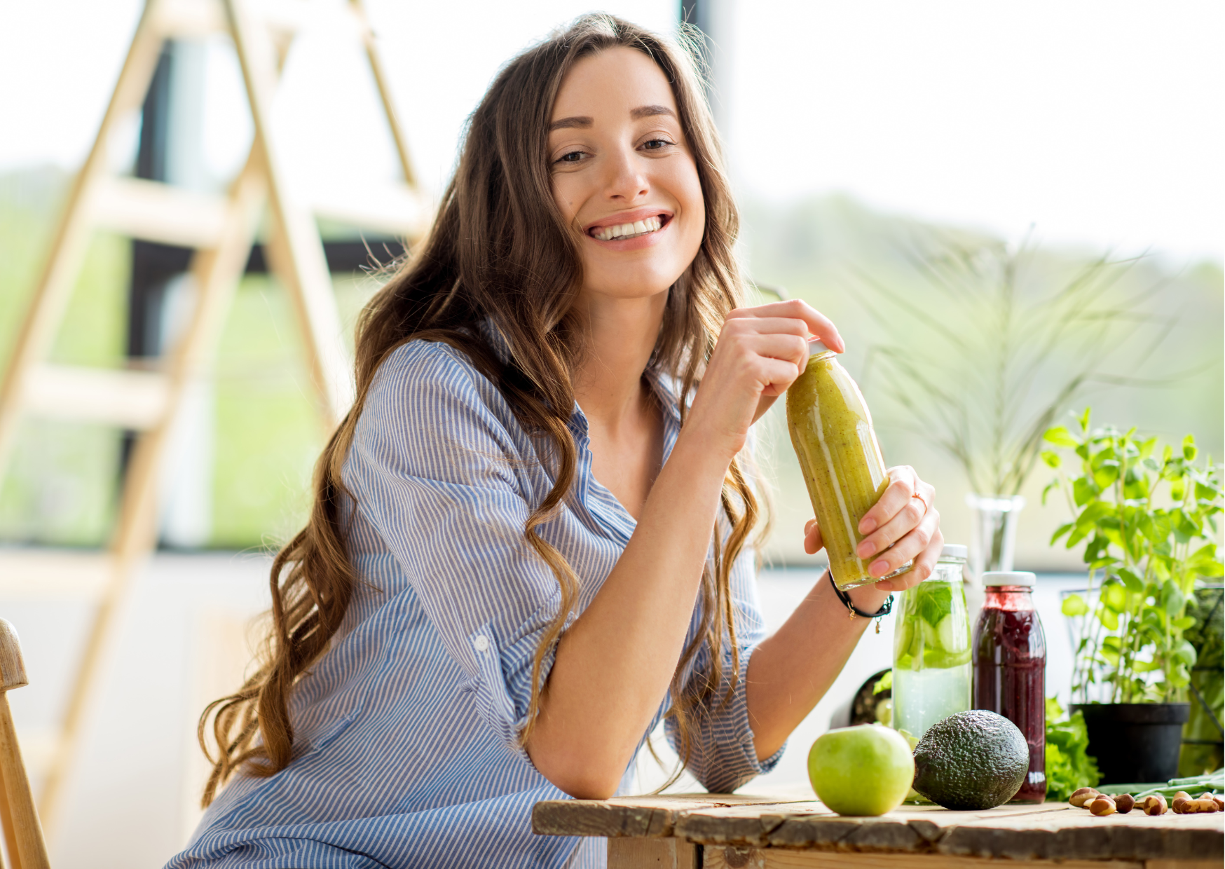 Em destaque está um mulher branca, sorrindo para a foto, sentada em frente à uma mesa de madeira, em ambiente amplo, claro e aberto, com plantas, ucos, frutas e castanhas sobre a mesa, vestindo uma camisa de mangas três quartos azul com listras brancas, cabelos longos até os quadris da cor castanho claro e segurando uma garrafa com um suco amarelo esverdeado.