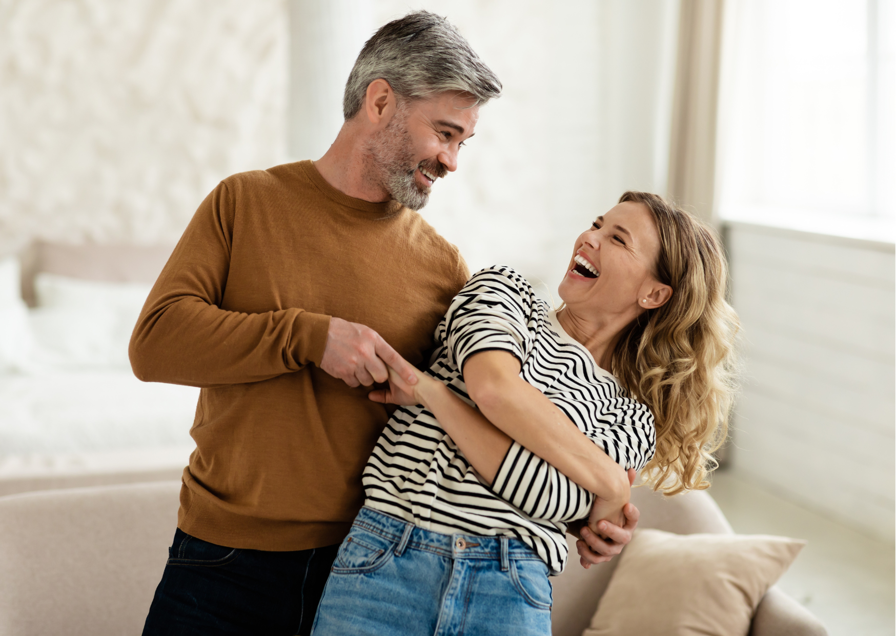 Em destaque, numa sala residencial ampla a e clara, está um casal branco de meia idade, vestindo ambos jeans, ele com uma camiseta de mangas longas na cor ocre e uma uma blusa de mangas 3/4 listrada de preto e branco, enlaçados como se estivessem dançando.