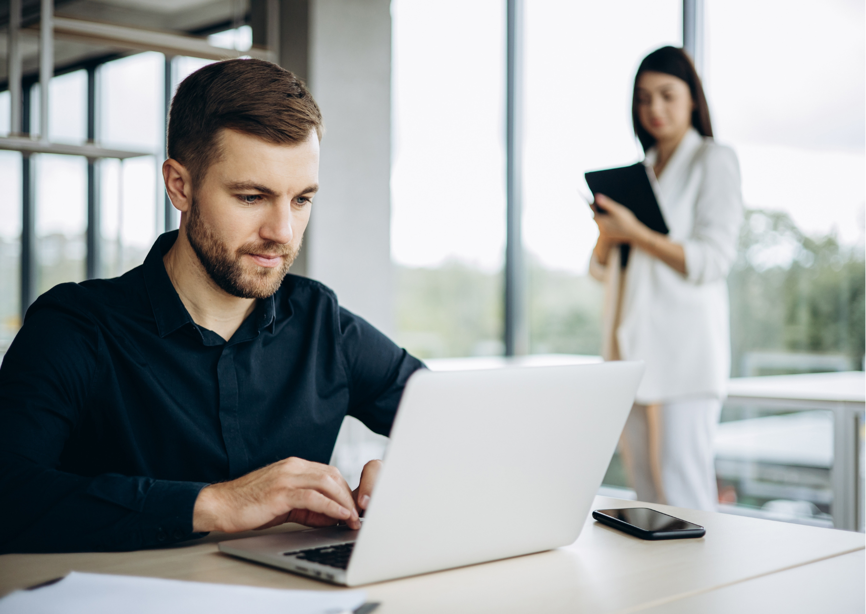 Em um ambiente claro, com paredes de vidro, a fotografia mostra em foco um homem branco, vestindo camisa social preta, sentado em frente à um notebook, focado na ação; ao fundo é possível ver uma mulher de pé, utilizando um caderno e vestindo roupas brancas.