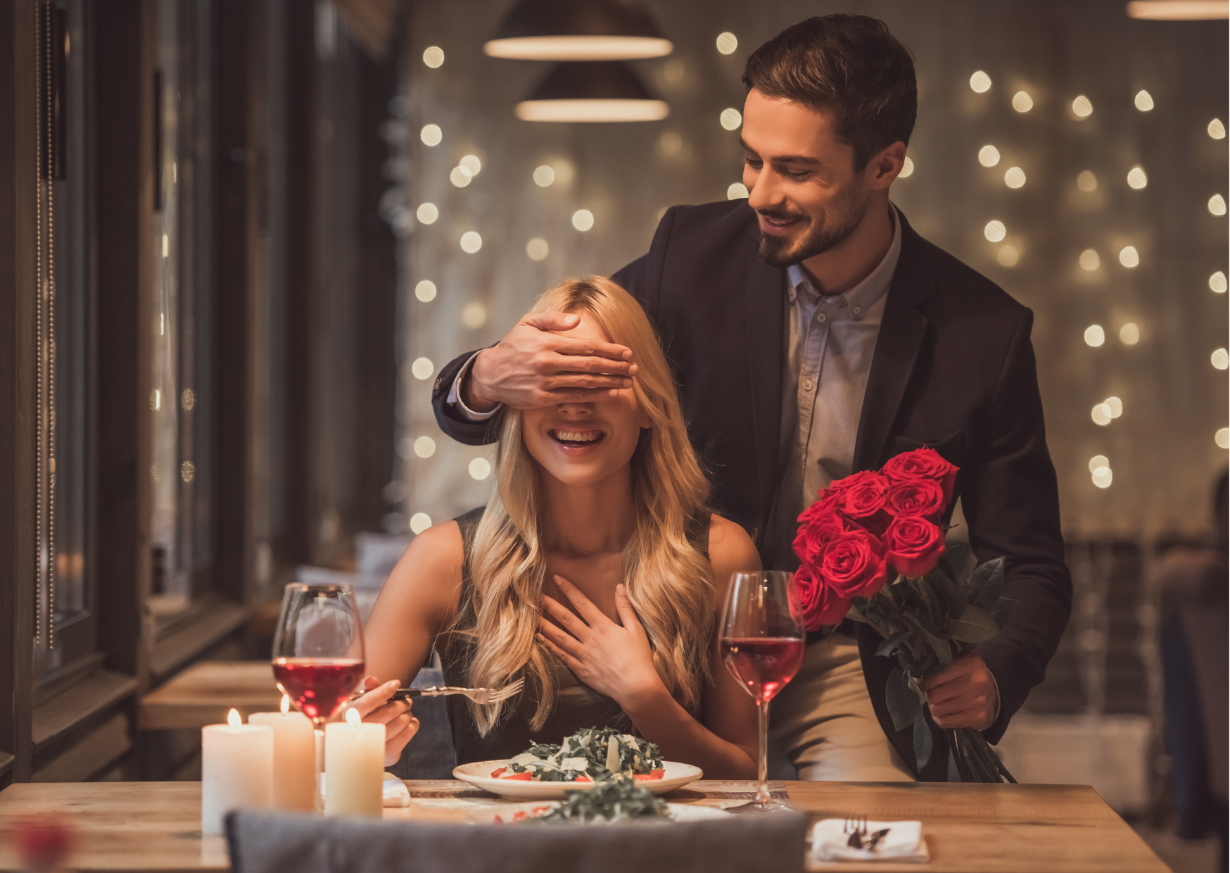 Em um ambiente romântico como um restaurante com cascata de luz ao fundo, uma mulher branca e loira está sentada na mesma posta, com os olhos tampados pela mão do homem que está parado atrás dela, surpreendendo-a com um buquê de rosas vermelhas, representando as ideias para presentes de dia dos namorados.