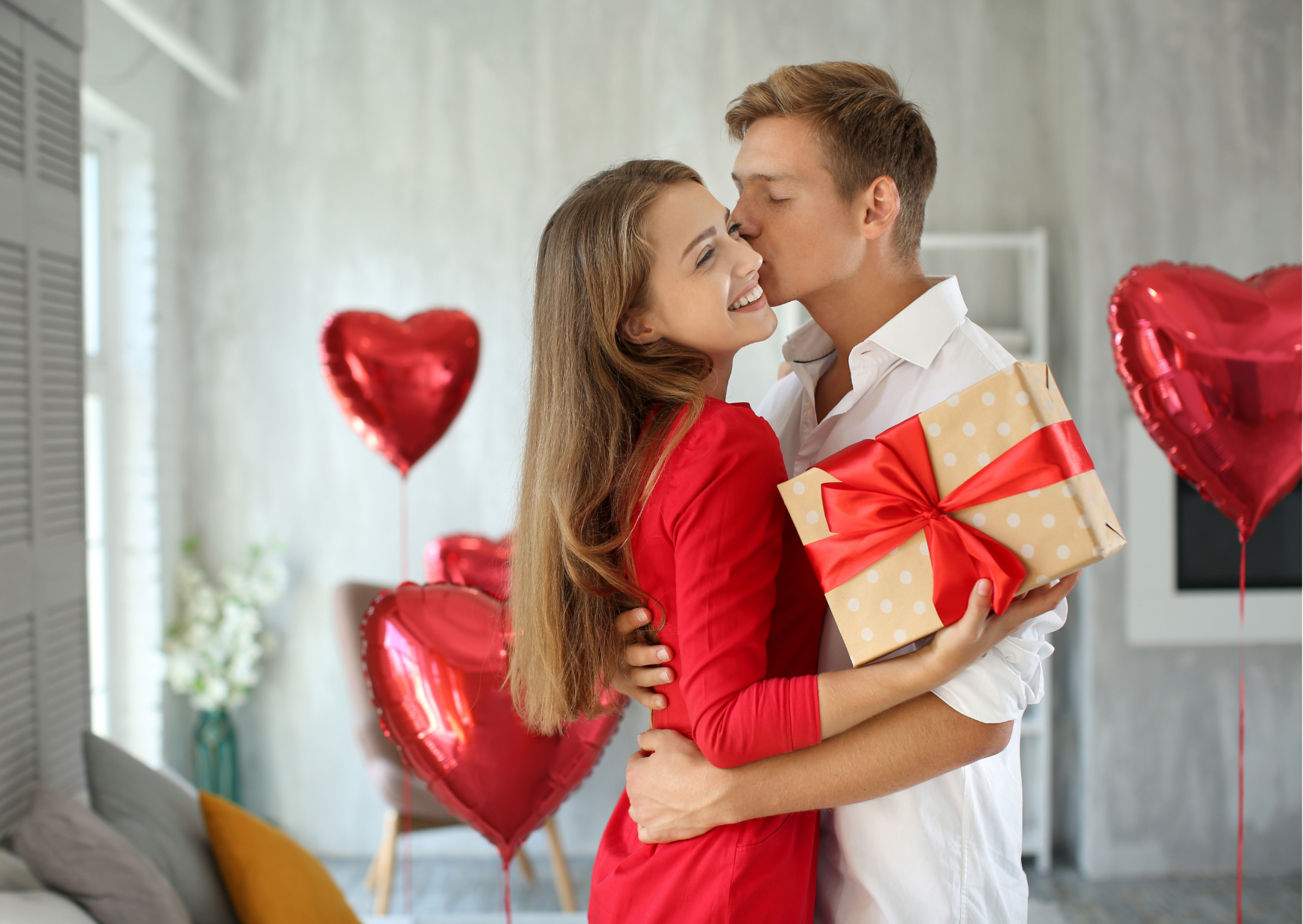 Representando as ideias de presentes para o Dia dos Namorados, está um jovem casal, ambos brancos e de cabelos claros e loiros, ela usando um vestido vermelho e segurando uma caixa de presente, ele usa camisa branca, ambos se abraçando e o rapaz dando um beijo na bochecha da moça,  em um ambiente semelhante à uma sala de estar, com balões vermelhos em formato de coração decorando.