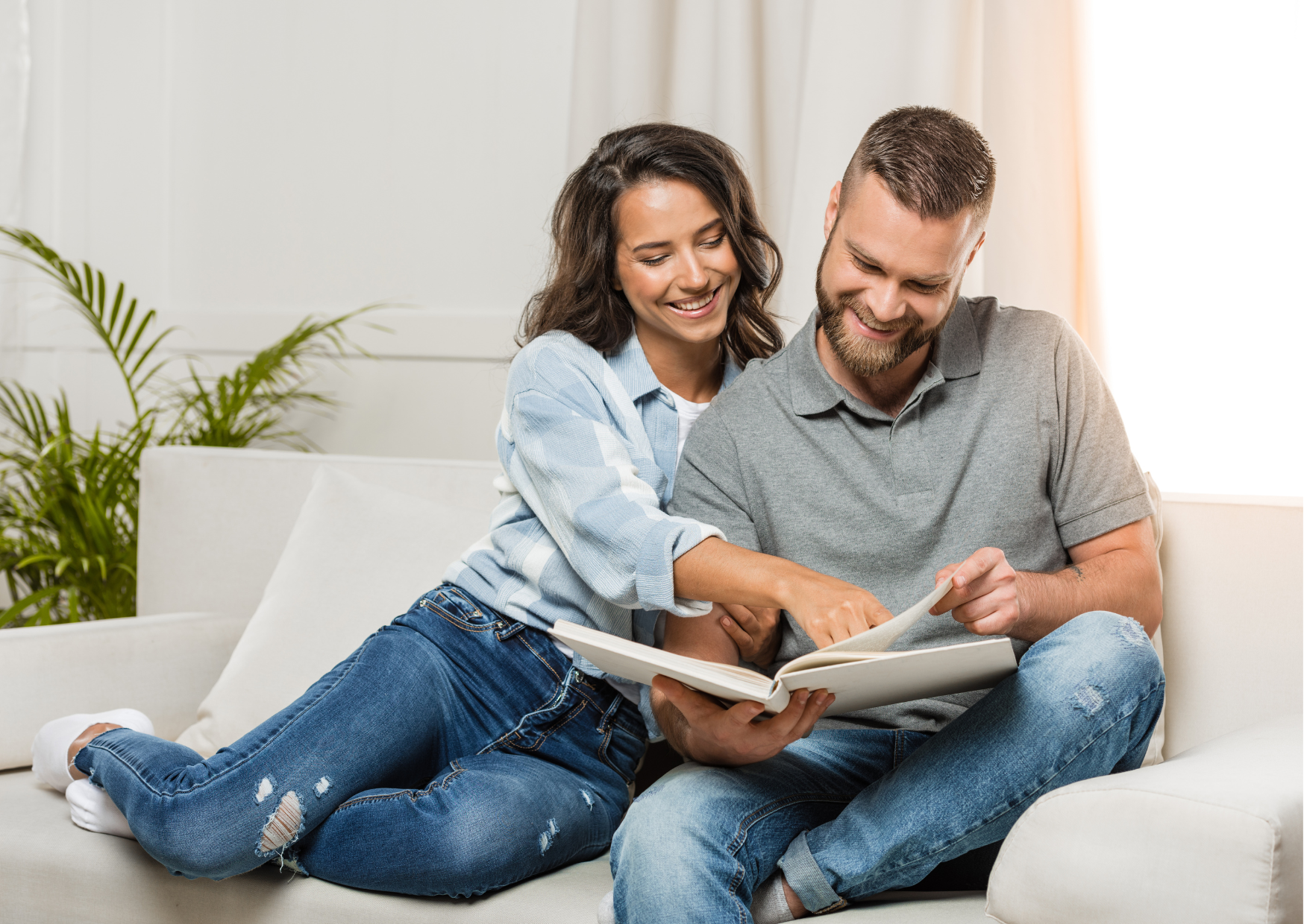 Representando as ideias de presentes para o Dia dos Namorados, a fotografia mostra um casal de pessoas brancas sentado em um sofá, ela vestindo calça e blusa jeans, ele vestindo calça jeans e blusa polo cinza e segurando um álbum de fotos no qual a mulher aponta um detalhe, ambos sorrindo.