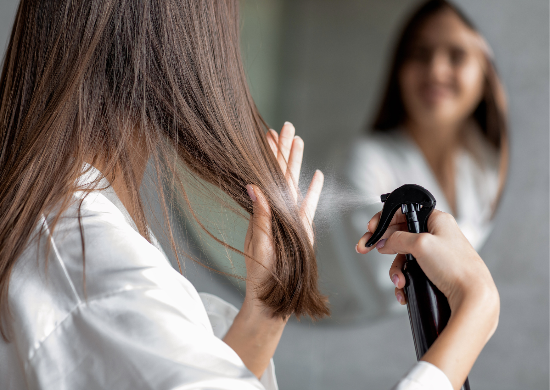 Representando como cuidar do cabelo em dias frios, está a foto de uma mulher branca, de costas, com cabelos castanhos e lisos, aplicando um spray em frasco preto nos fios, em frente ao espelho, do qual vemos apenas sua imagem refletida desfocada.