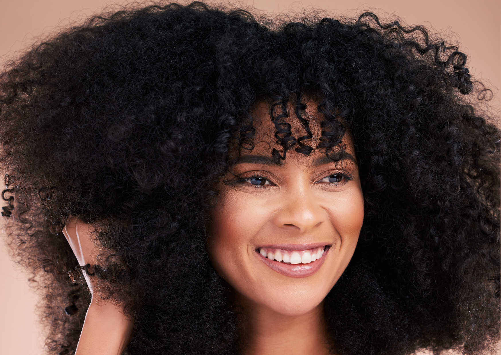 Representando as tendências de cores e corte de cabelo no outono-inverno 2023, a fotografia mostra uma mulher negro, com cabelos crespos com alto volume e corte com muitas camadas, sorrindo e com uma das mãos segurando mechas dos cabelos.