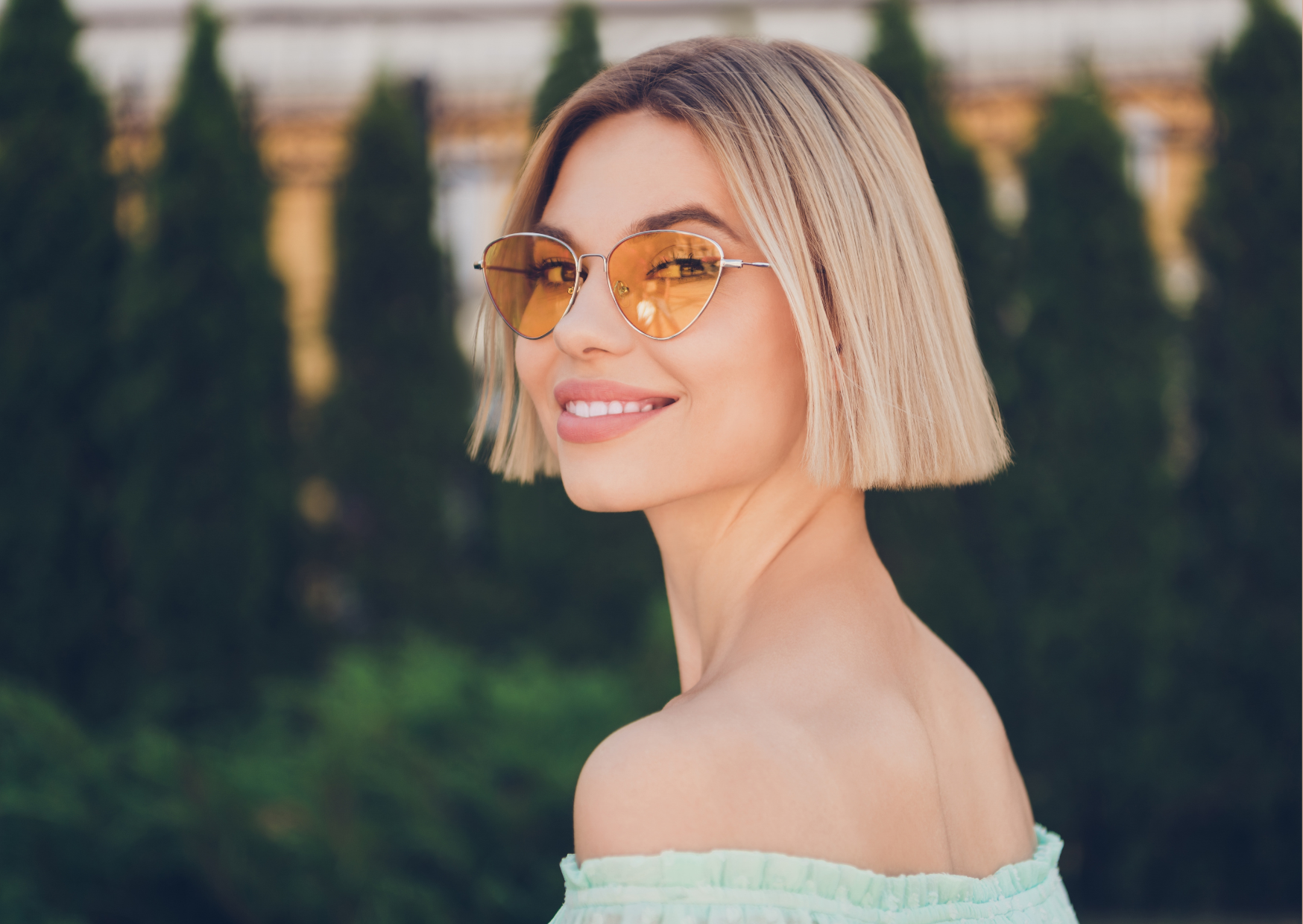 Fotografia ao ar livre, ao fundo desfocado estão árvores escuras e verdes, está uma mulher branca, com corte de cabelo blunt bob, fios lisos e loiros platinados na altura do queixo, sorrindo para a foto e usando óculos com lentes amareladas.