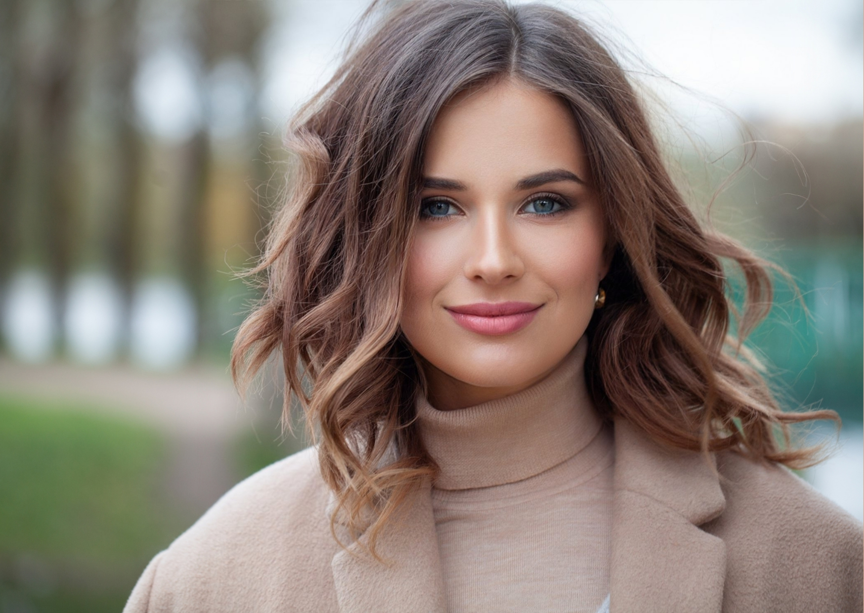 Representando as tendências de cores e corte de cabelo no outono-inverno 2023, a fotografia mostra uma mulher branca, com cabelos louro escuro, cortados na altura dos ombros, estilo long-bob, vestindo roupas em tons bege e areia, sorrindo com lábios fechados.