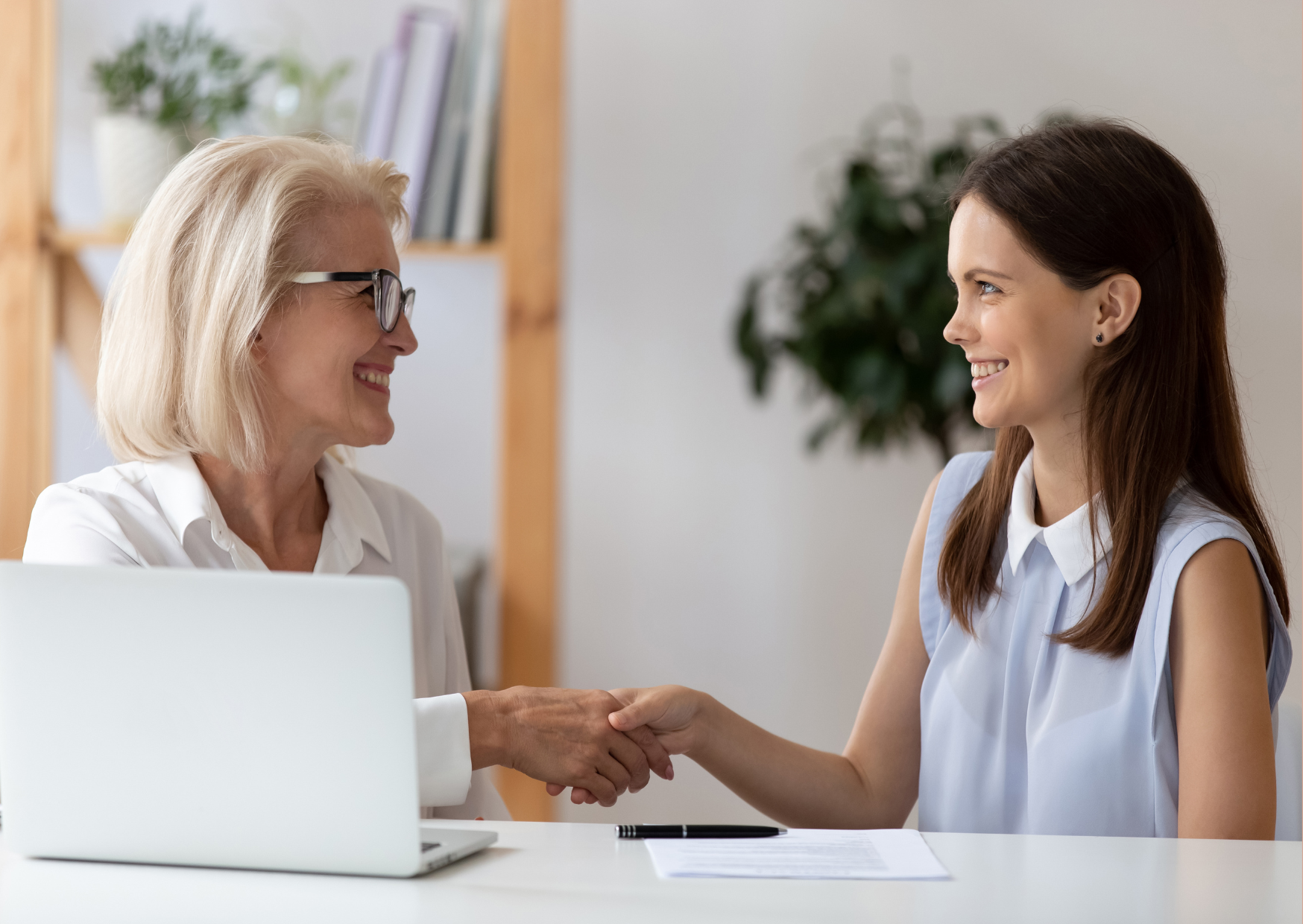 Exemplificando o pós-venda para distribuidores, a fotografia mostra duas mulheres sentadas em frente a uma mesa com notebook, apertando a mão uma da outra e sorrindo; à esquerda, mulher branca, idosa, usa óculos e camisa branca; à direta mulher branca, jovem, usa camisa sem mangas azul clara.